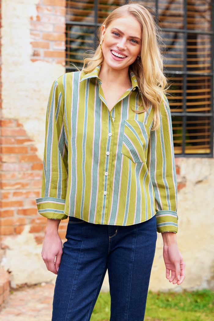 Woman wearing a green and yellow striped shirt with blue jeans standing in front of a brick wall.