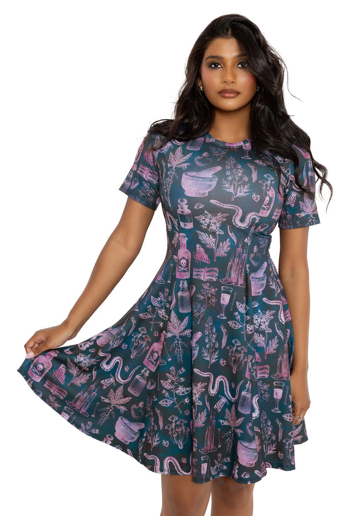 Longline witchy tee dress with apothecary bottle, herb and potion artwork in moody pink and teal tones. This gothic skater dress blends whimsigoth, dark fairycore and cottage witch fashion with a comfy longline silhouette perfect for alternative goth and fantasy witch styling.