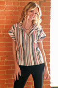 A woman wearing a short-sleeve striped button-up shirt with vertical stripes in beige, red, blue, and gray, paired with black pants, standing against a brick wall.
