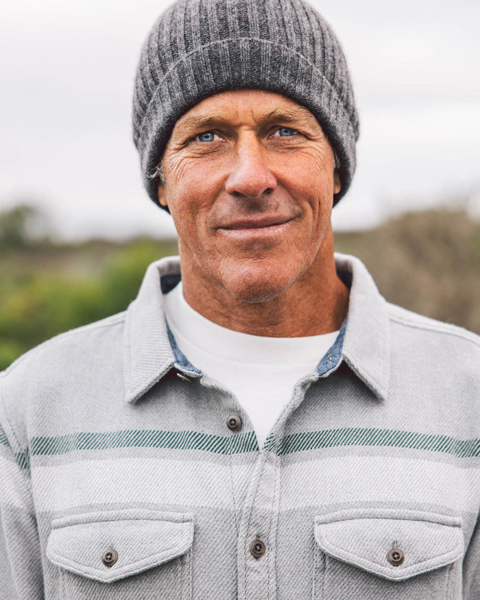 Man wearing a gray knit beanie and light gray jacket with a striped shirt underneath, standing outdoors.