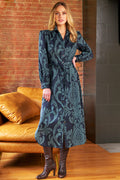 Woman wearing a long, patterned dress standing in a room with a brick wall and brown couch.