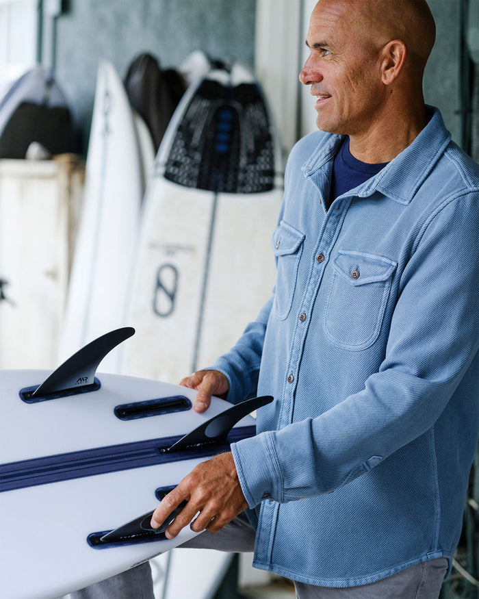 Kelly Slater adjusting fins on a surfboard in a workshop setting