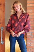 Woman wearing a red and brown patterned blouse with blue jeans standing against a brick wall.