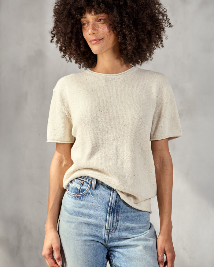 A woman wearing a beige knit top with short sleeves and a round neckline, paired with blue jeans.
