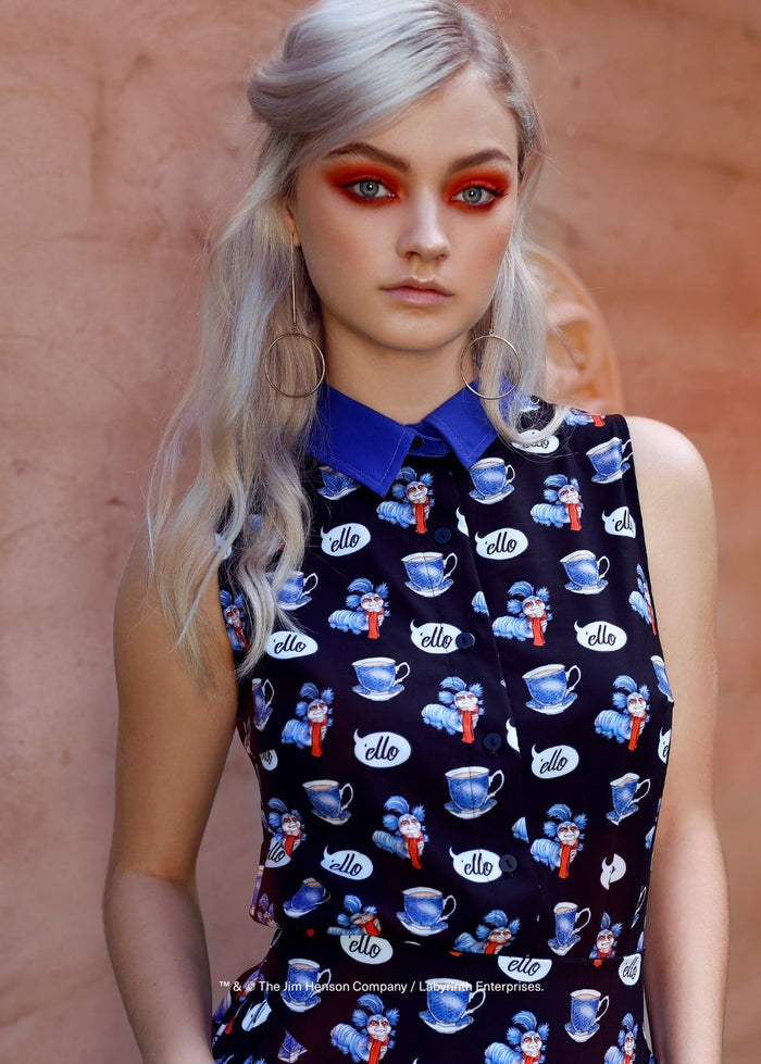 A model with platinum blonde hair and dramatic red eye makeup wears a sleeveless navy dress with a blue collar and a repeating pattern of blue teacups and white speech bubbles containing the text 'ello'. She is posed against a textured, warm terracotta-colored wall.