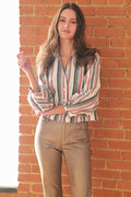A woman with long wavy brown hair stands against a red brick wall wearing a long-sleeve button-up shirt with vertical stripes in cream, red, and gray, paired with tan pants.