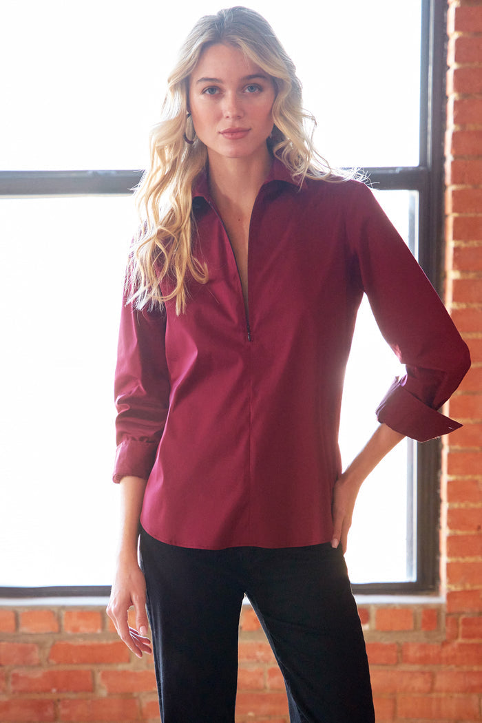 A woman modeling a long-sleeve button-up shirt in deep burgundy, paired with black pants, standing in front of a brick wall and window.