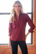 A woman modeling a long-sleeve button-up shirt in deep burgundy, paired with black pants, standing in front of a brick wall and window.
