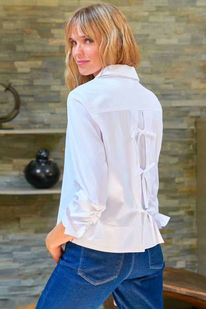 Woman wearing a white blouse with sheer panels and blue jeans against a stone wall.