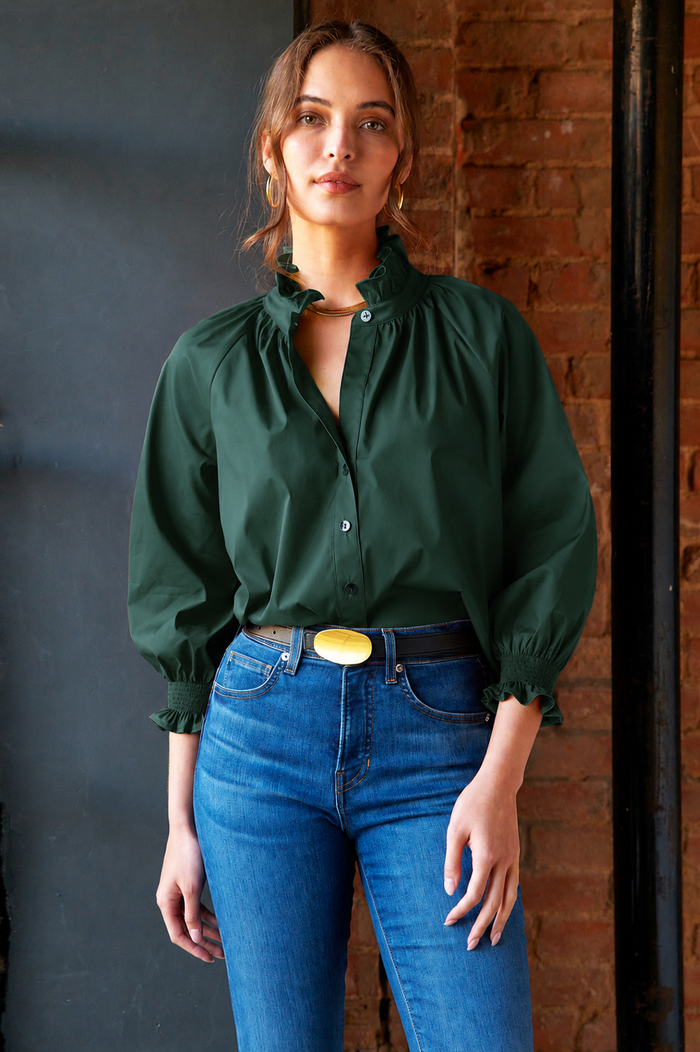 A woman wearing a dark green long-sleeve blouse with a ruffled high collar and gathered cuffs, paired with blue jeans and a yellow belt, standing against a dark gray wall and brick wall background.