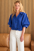 A woman wearing a vibrant blue crinkled short-sleeve button-up shirt and white trousers, standing in front of a brown leather sofa and a wooden slat wall.