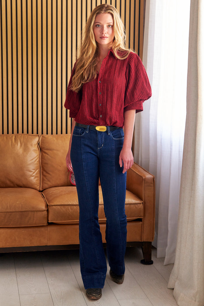 A woman modeling a red puffed-sleeve button-up shirt and dark blue jeans with a yellow belt, standing in front of a brown leather sofa and a slatted wood wall.