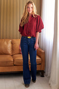 A woman modeling a red puffed-sleeve button-up shirt and dark blue jeans with a yellow belt, standing in front of a brown leather sofa and a slatted wood wall.