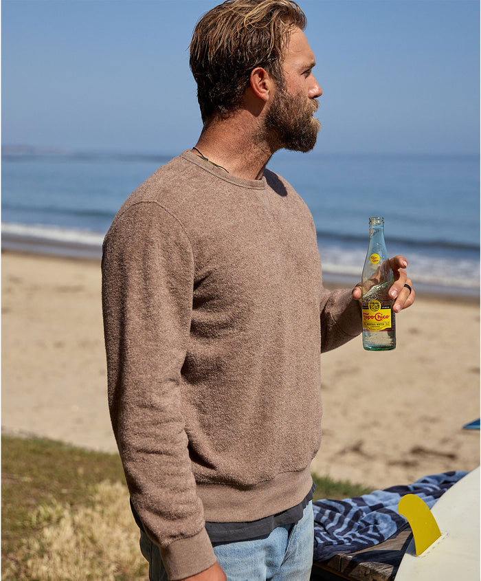 Man wearing brown sweater holding a bottle on a beach with ocean in the background
