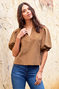 Woman wearing a brown blouse and blue jeans against a stone wall.