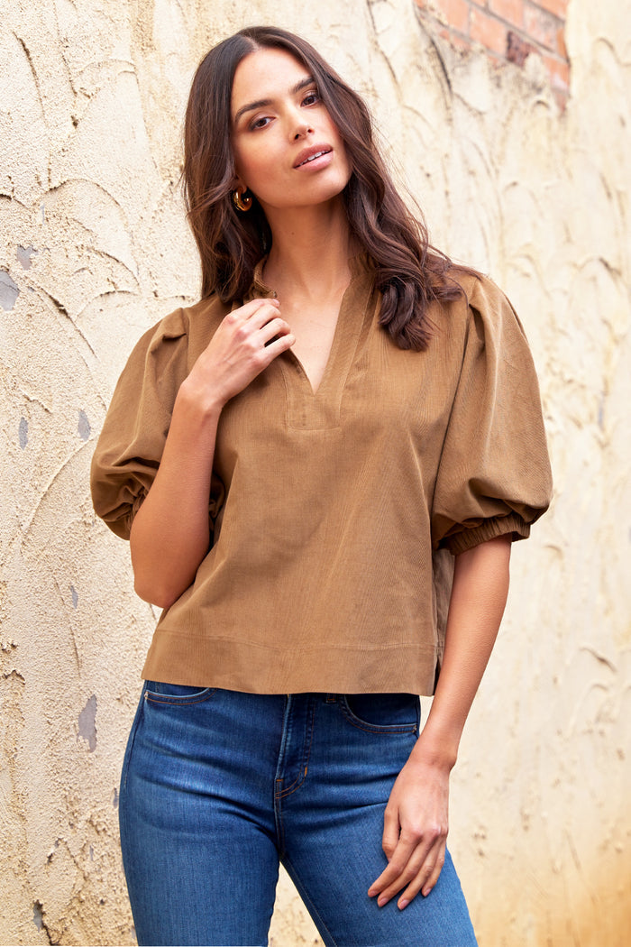 Woman wearing a brown blouse and blue jeans against a stone wall.
