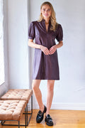 Woman wearing a purple dress standing in a room with wooden floors and a white wall.