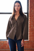 A woman modeling a dark brown button-up shirt with a split V-neck and long sleeves, paired with dark blue jeans, standing in front of a brick wall and window.