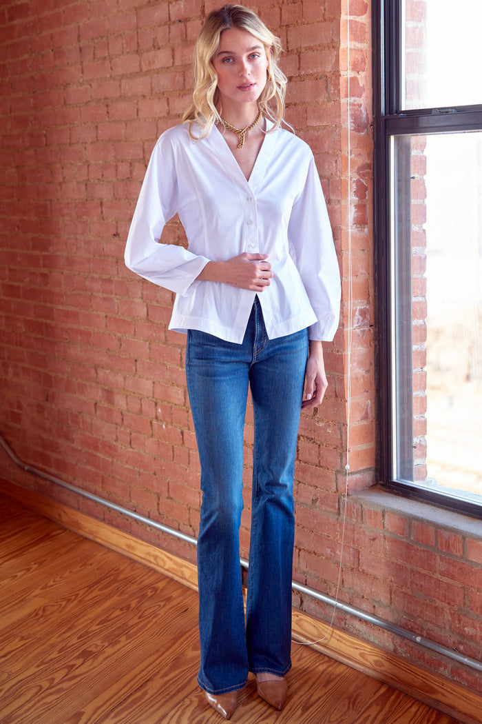 Woman wearing a white blouse and blue jeans standing in front of a brick wall.