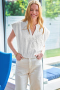 A woman wearing a white textured jacquard popover top with cap sleeves and a V-neckline, paired with light-colored trousers.
