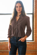 A woman wearing a dark brown long-sleeve button-up shirt and dark blue jeans, standing in front of a brick wall and window.