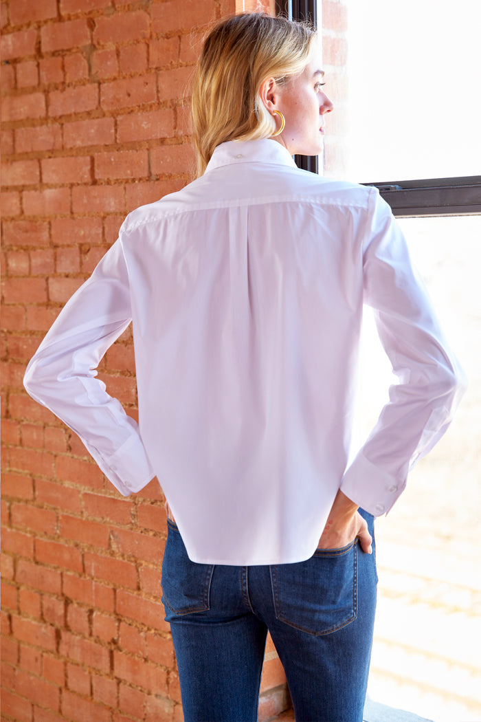 A woman wearing a white long-sleeve button-down shirt and blue jeans, shown from the back in front of a brick wall and window.