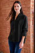 A woman wearing a black button-up shirt and blue jeans stands against a red brick wall.