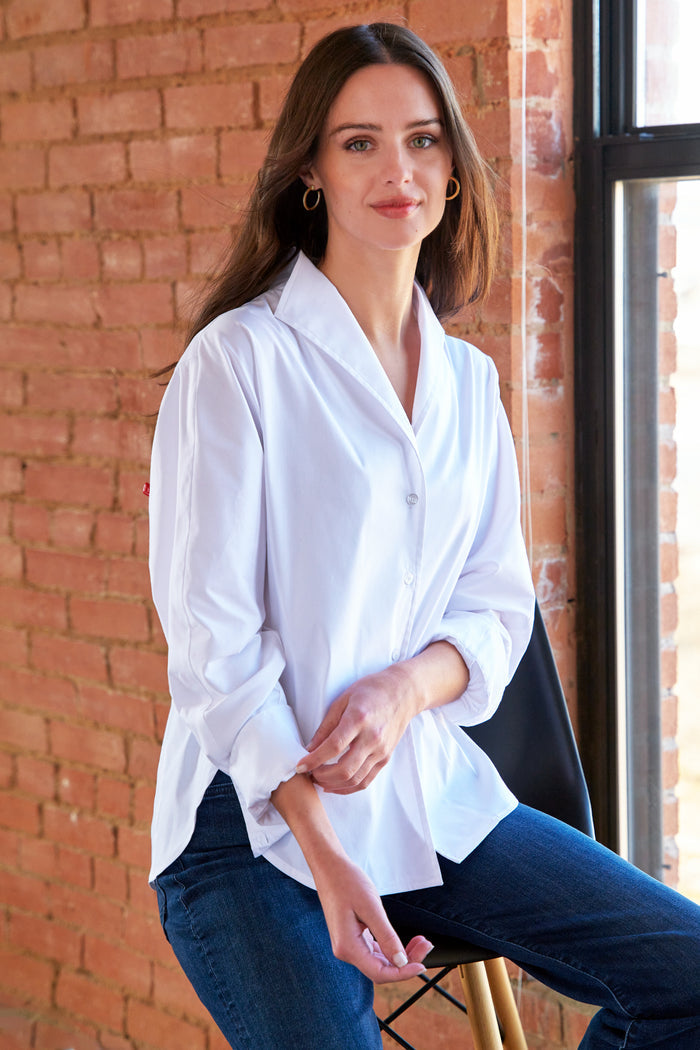 Woman wearing a white blouse and blue jeans sitting against a brick wall.