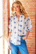 Woman wearing a white blouse with blue floral patterns and blue jeans against a brick wall.