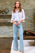 Woman wearing a white blouse and light blue jeans standing in a room with stone walls and wooden furniture.