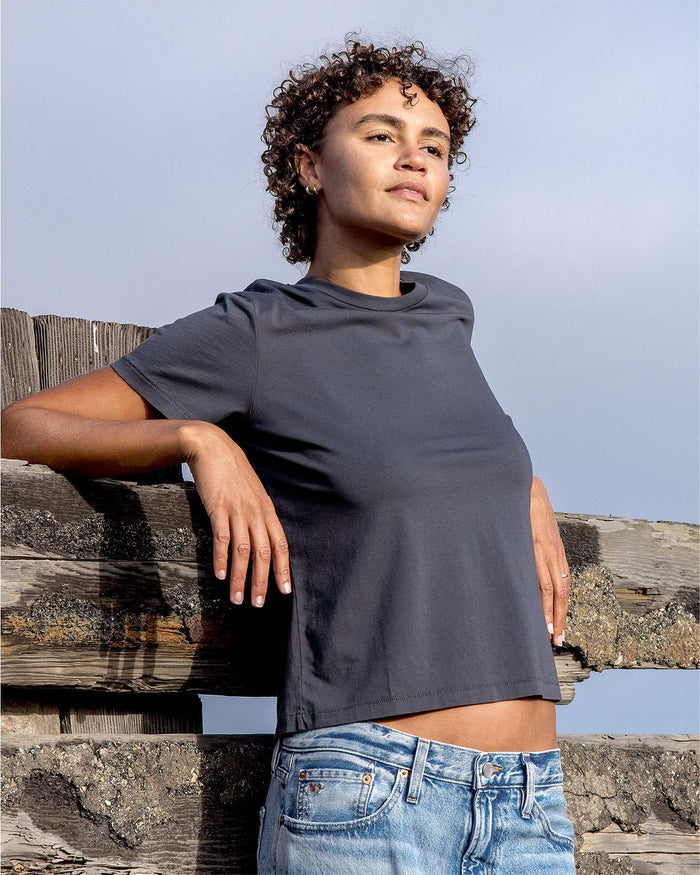 Person wearing a dark gray t-shirt and blue jeans leaning against a wooden fence with a clear sky background.