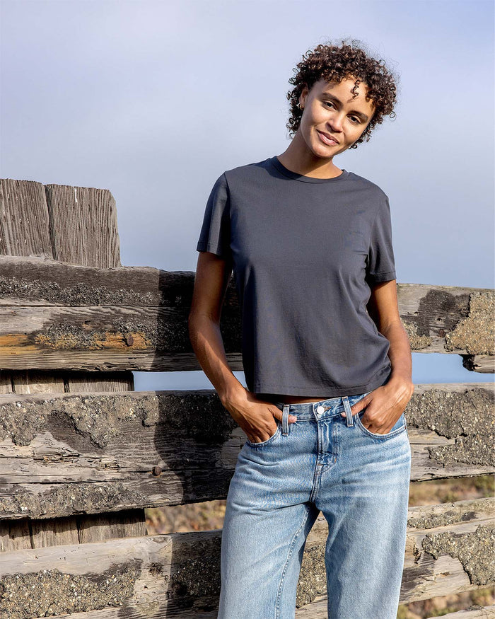 Person wearing a dark gray t-shirt and light blue jeans standing on wooden planks with a clear sky background