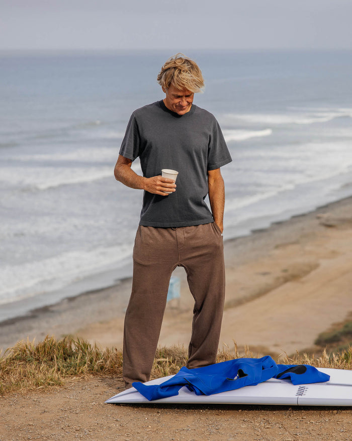 Man wearing brown sweats standing on a beach holding a coffee cup with a surfboard and blue jacket nearby.