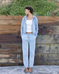 Woman wearing a light blue hoodie and pants standing against a wooden wall.