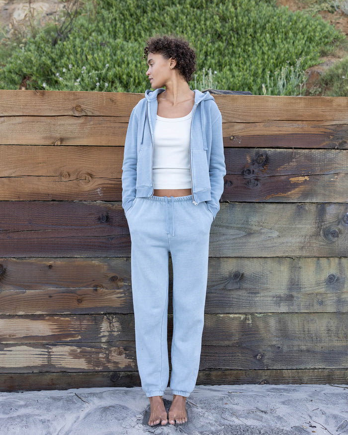 Woman wearing a light blue hoodie and pants standing against a wooden wall.