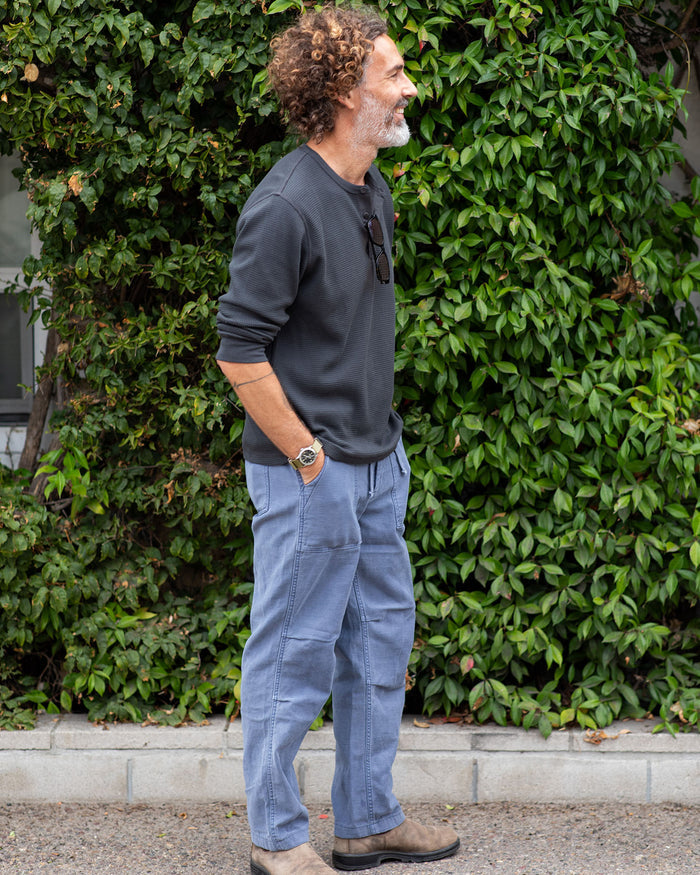 Man wearing a dark shirt and blue pants standing in front of a green bush.