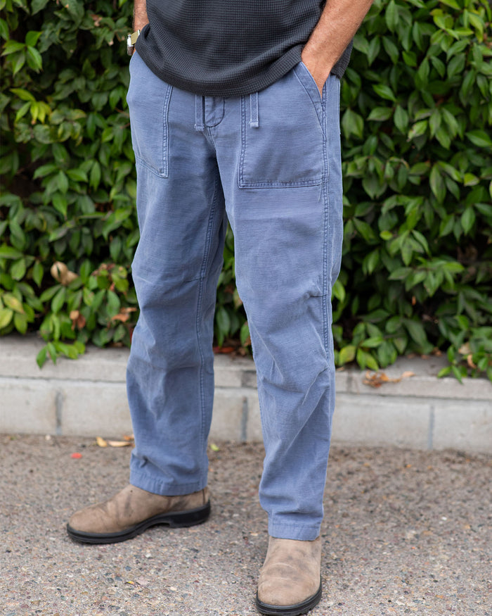 Person wearing blue pants and brown shoes standing in front of green foliage.