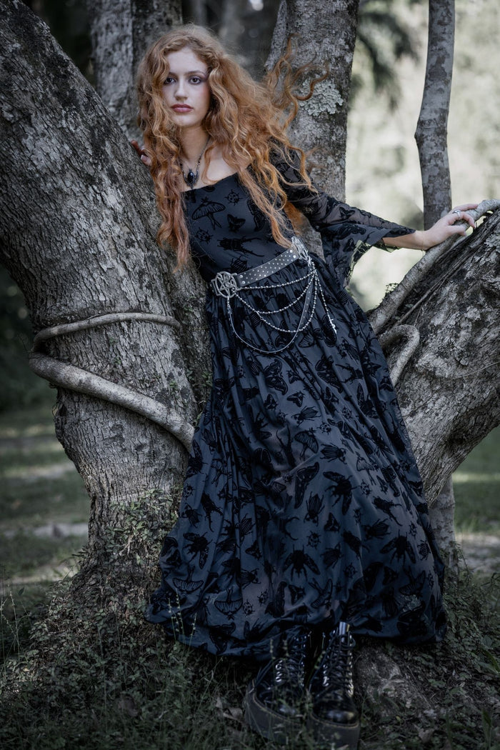 Black burned velvet maxi dress featuring Ami Raithe moth artwork with fluttering insects scattered across sheer mesh fabric. This whimsigoth maxi dress blends dark fairycore, gothic fashion and fantasy witch aesthetics with dramatic flute sleeves and a romantic flowing silhouette.