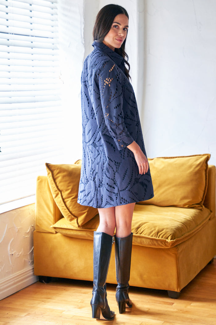 Woman wearing a blue knitted dress and black knee-high boots standing in a room with a yellow couch.