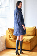 Woman wearing a blue knitted dress and black knee-high boots standing in a room with a yellow couch.