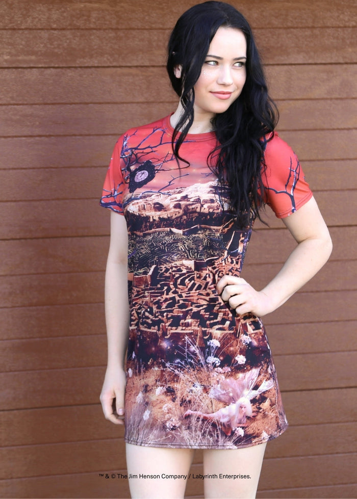 A woman modeling a short-sleeve t-shirt dress with a red crew neck and a detailed western landscape print in brown, beige, and black, posed against a brown wooden wall.