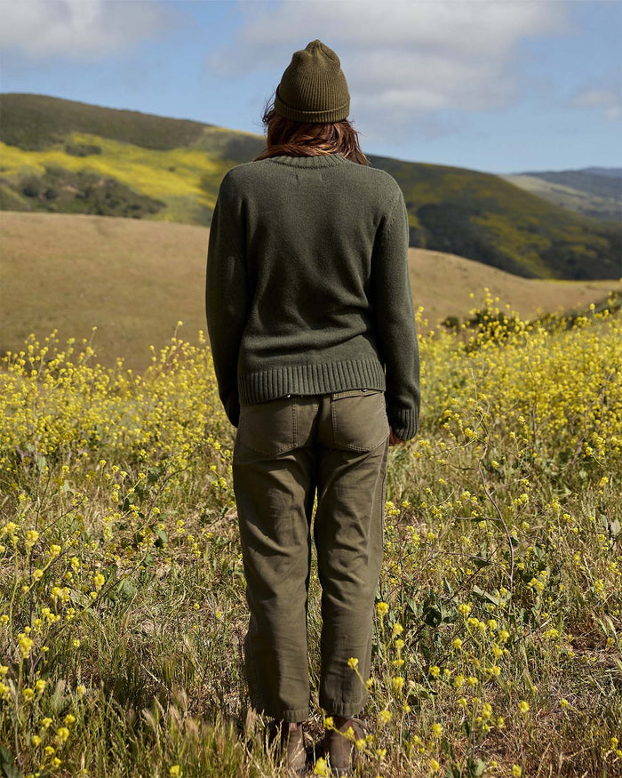 Person wearing a green sweater and beanie standing in a field of yellow flowers with rolling hills in the background.