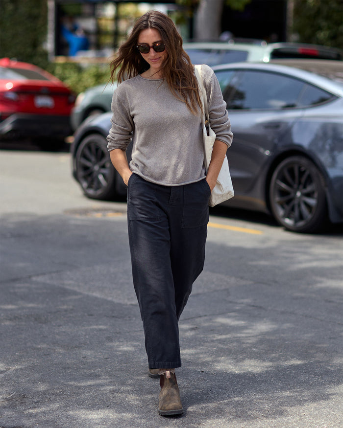 Woman walking on a street wearing a gray sweater and dark pants with a white bag.