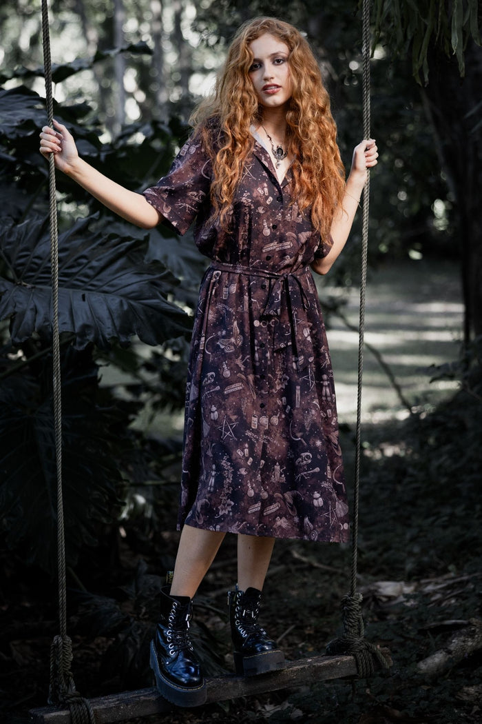 Witchy button-through midi dress featuring Ami Raithe artwork with potion bottles, bones, butterflies and mystical apothecary details scattered across a rich earthy print. This whimsigoth dress blends cottage witch, dark academia and fantasy witch fashion with a floaty A-line silhouette and vintage-inspired styling.