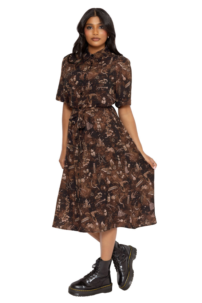 Alternative witchcore shirt dress with Ami Raithe potion artwork, occult details and dark botanical elements inspired by gothic apothecary aesthetics. Featuring a button-up bodice, tie waist and flowing midi skirt, this fantasy witch dress is perfect for cottage witch and modern pagan styling.