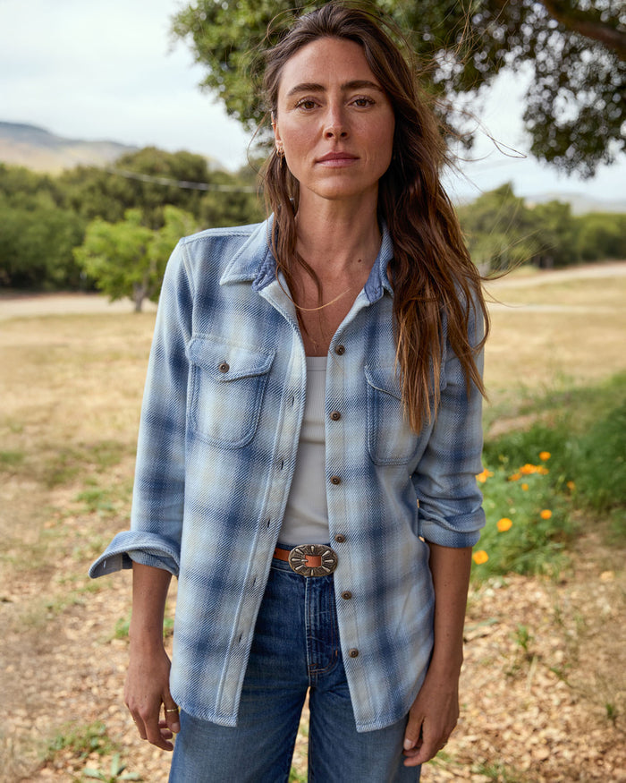 Woman wearing a blue plaid shirt and jeans standing outdoors with trees and grass in the background