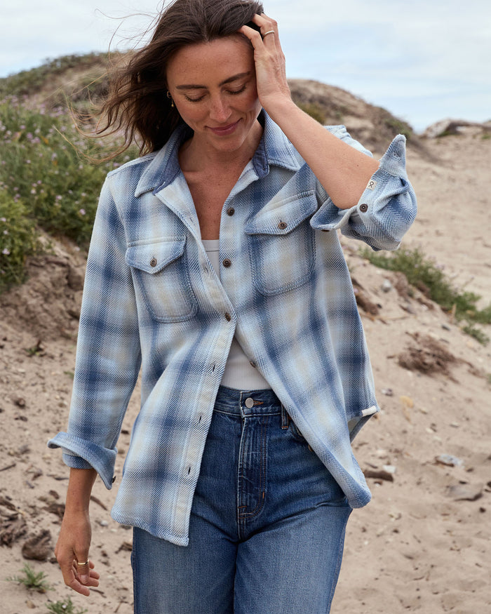 Woman wearing a blue plaid shirt and jeans standing on a sandy beach.