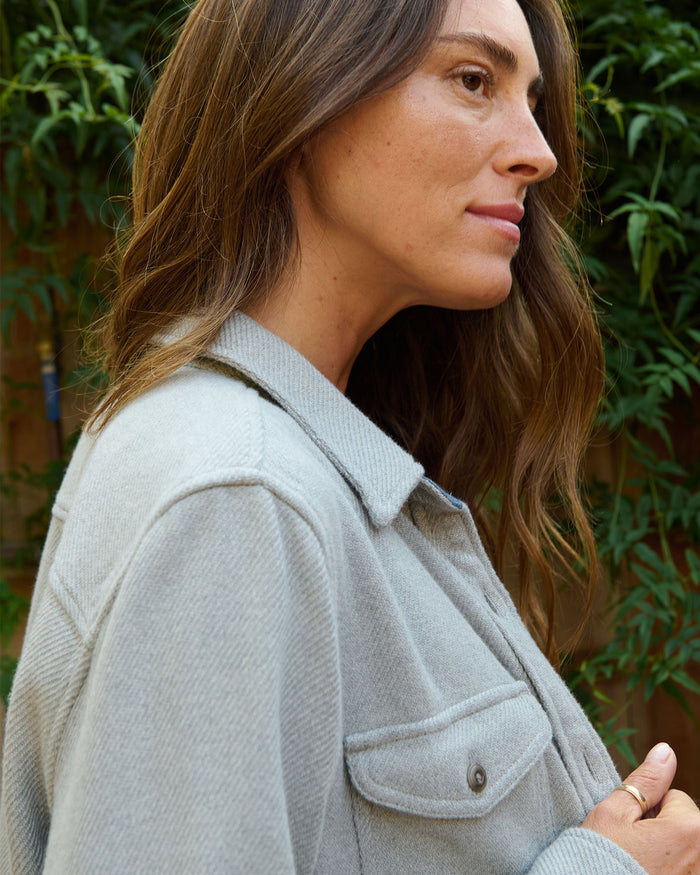 Woman wearing a light gray jacket outdoors with greenery in the background