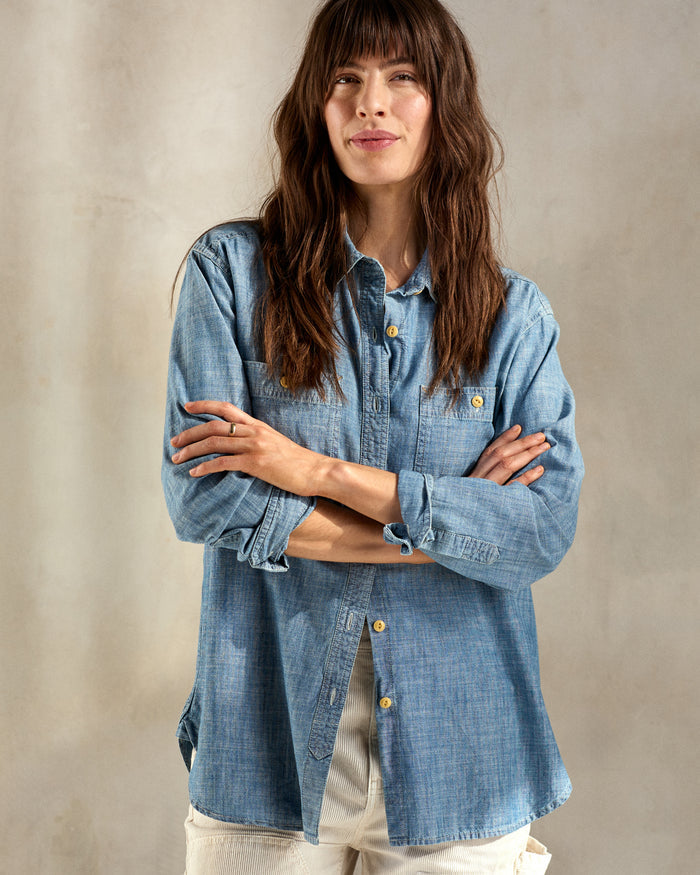 A woman wearing a long-sleeved, blue chambray utility shirt with button-up front and chest pockets, paired with light-colored pants.