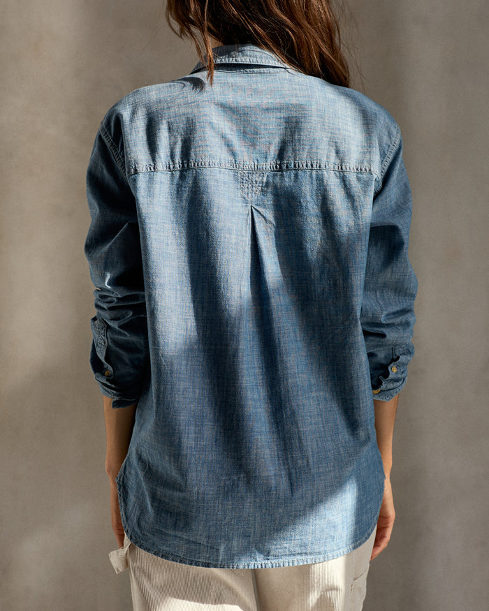 Back view of a woman wearing a long-sleeved, blue chambray utility shirt with button-up front and chest pockets, paired with light-colored pants.
